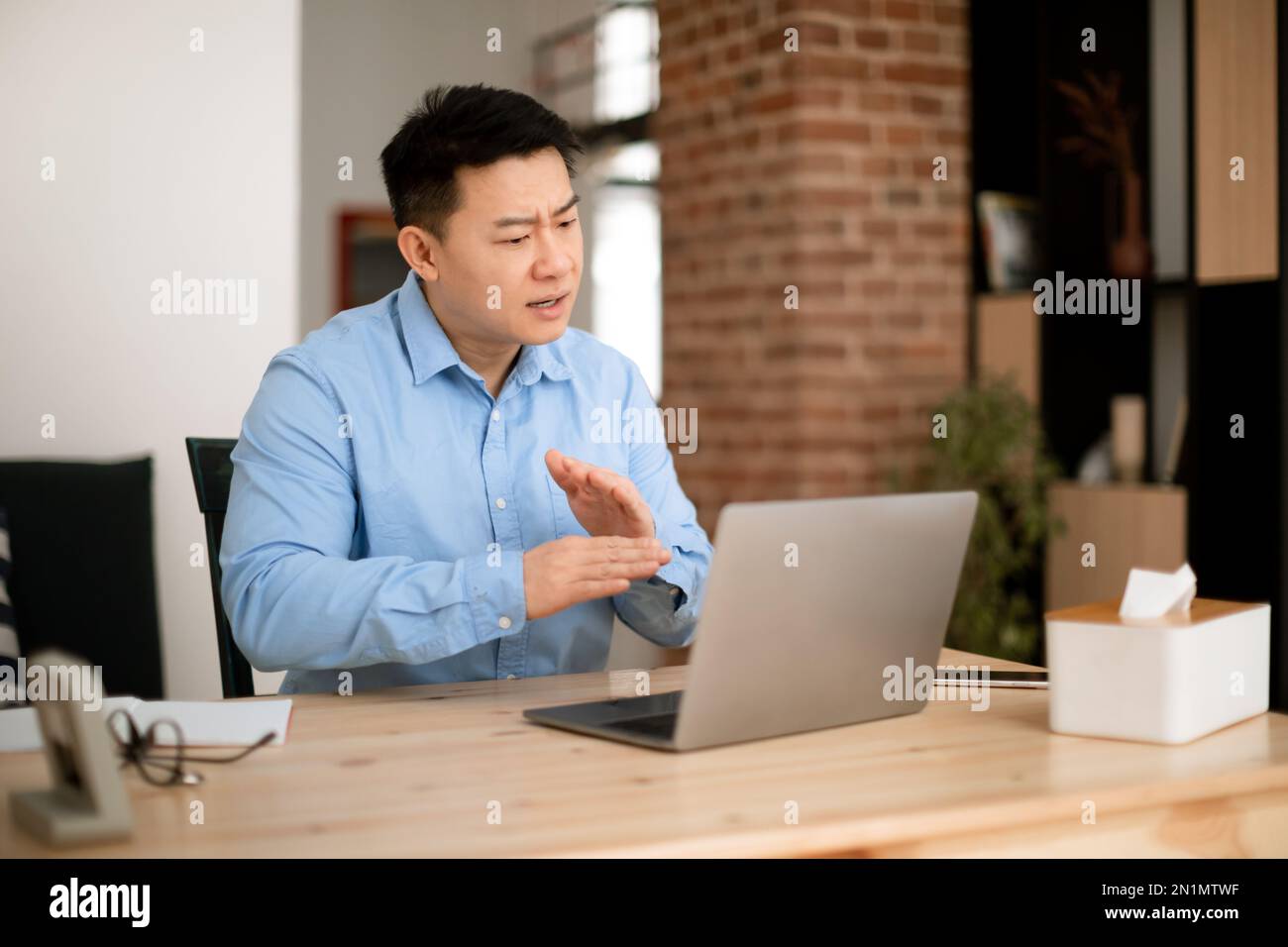 Homme d'affaires asiatique aux émotions assis à un bureau avec un ordinateur portable, se faisant des gestes tout en ayant des appels vidéo avec des collègues, espace libre Banque D'Images