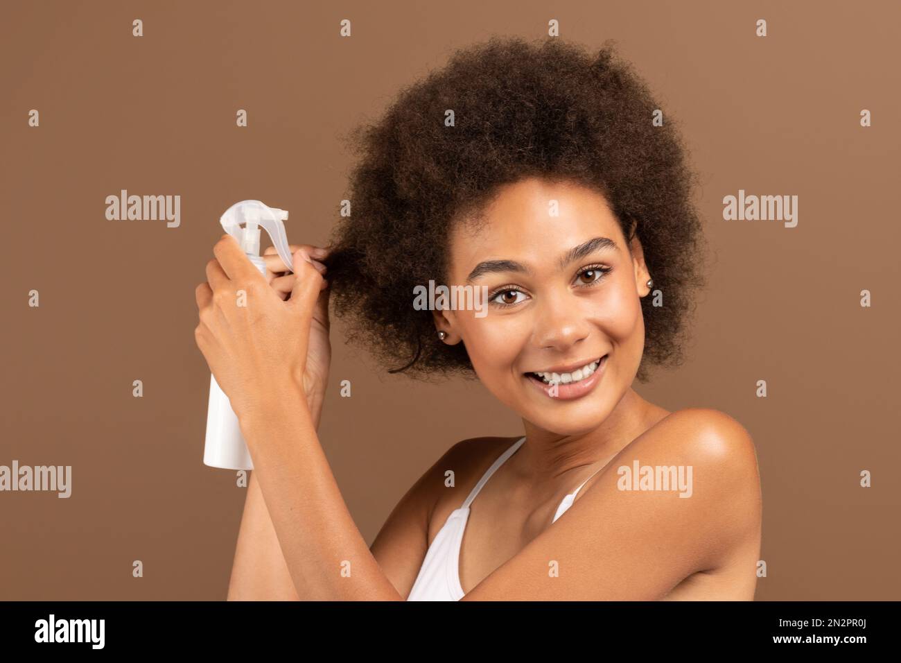 Souriante jeune afro-américaine curly femme avec une peau parfaite dans le haut blanc appliquant un spray sur ses cheveux Banque D'Images