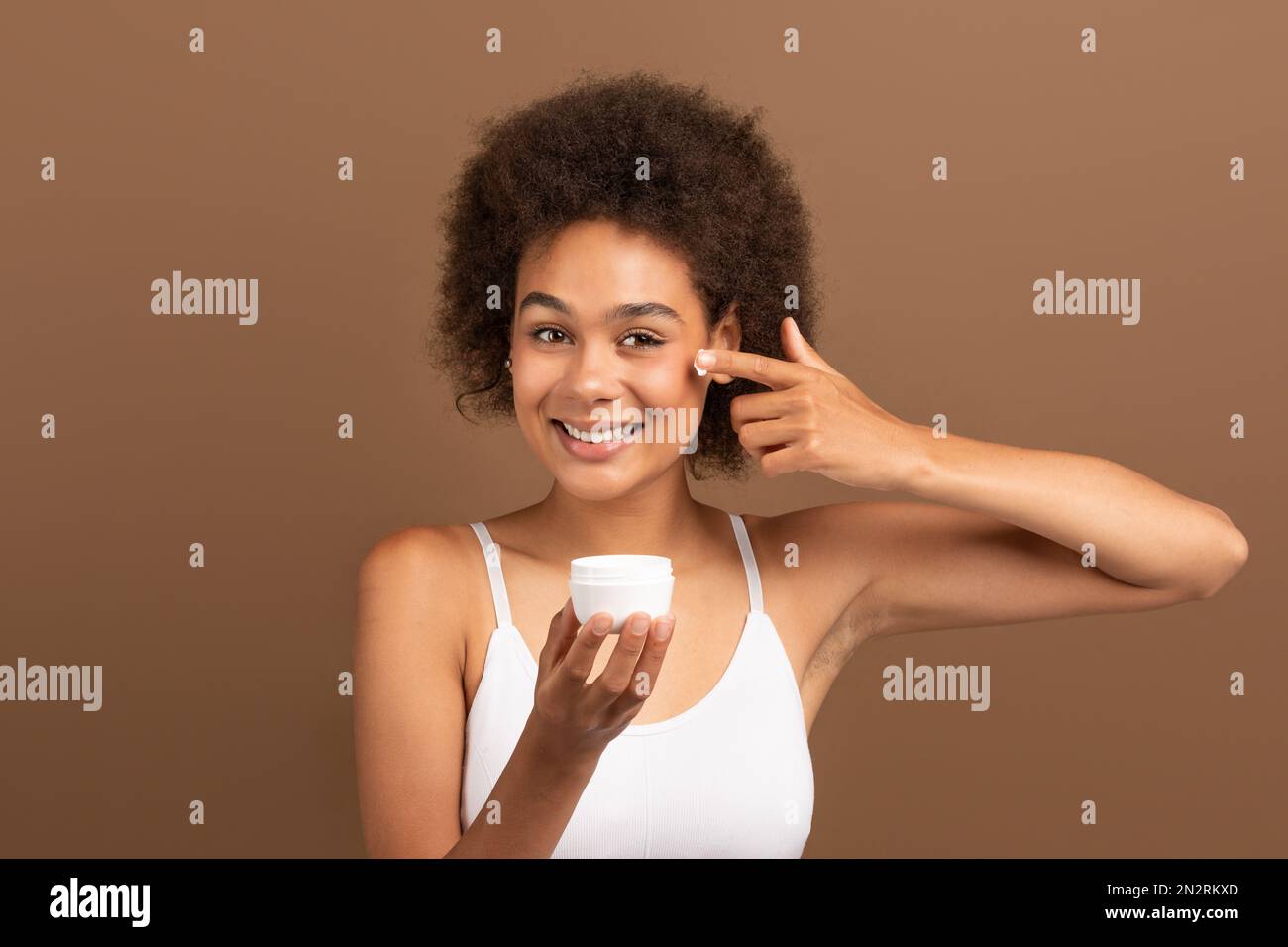 Gaie jeune afro-américaine curly femelle avec la peau parfaite dans le pot blanc de maintien du haut, appliquer la crème sur le visage Banque D'Images