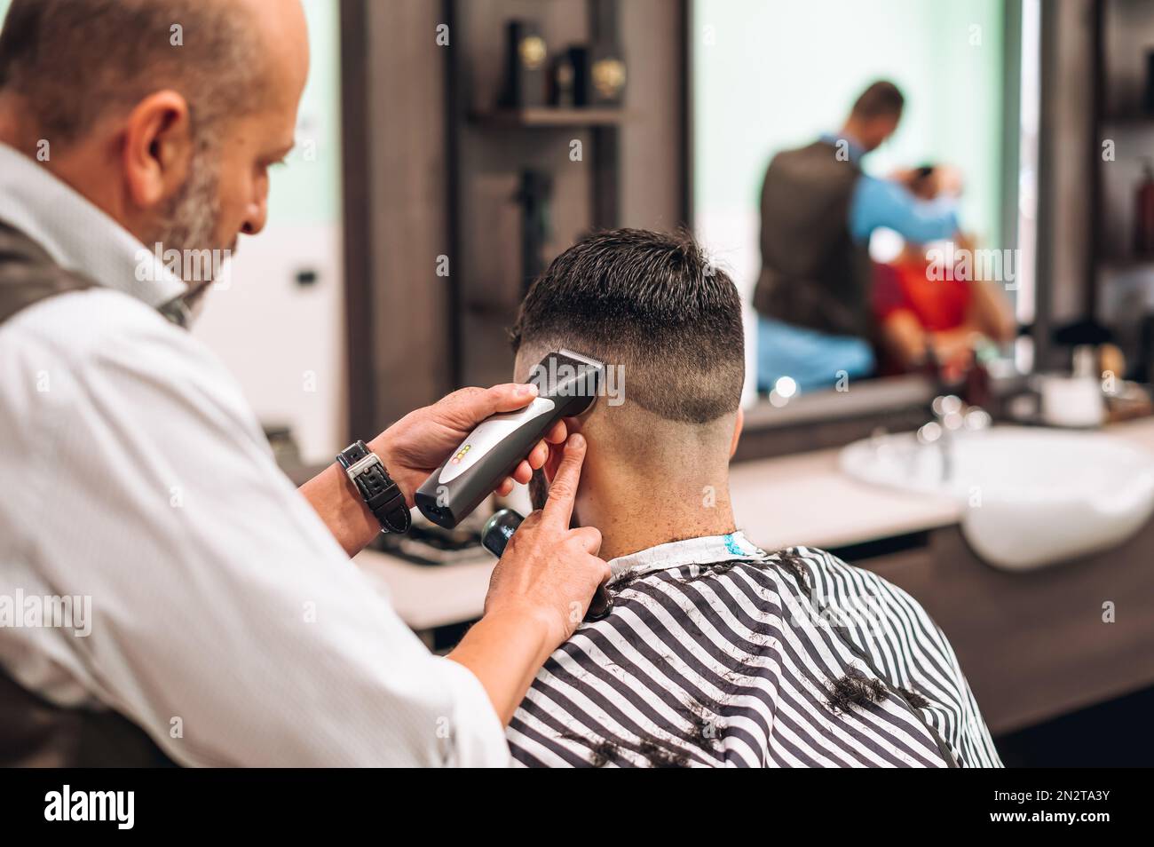 Vue arrière du crop barber tailler les cheveux sombres de la cliente dans le barbershop moderne pendant la journée de travail Banque D'Images