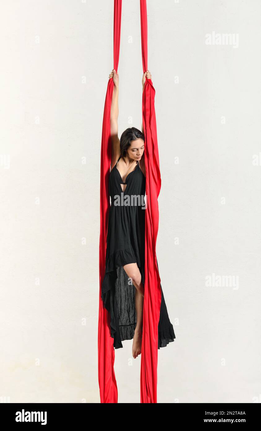 Gymnaste femelle pieds nus en robe noire suspendue sur des silks aériens rouges pendant les performances sur fond blanc Banque D'Images