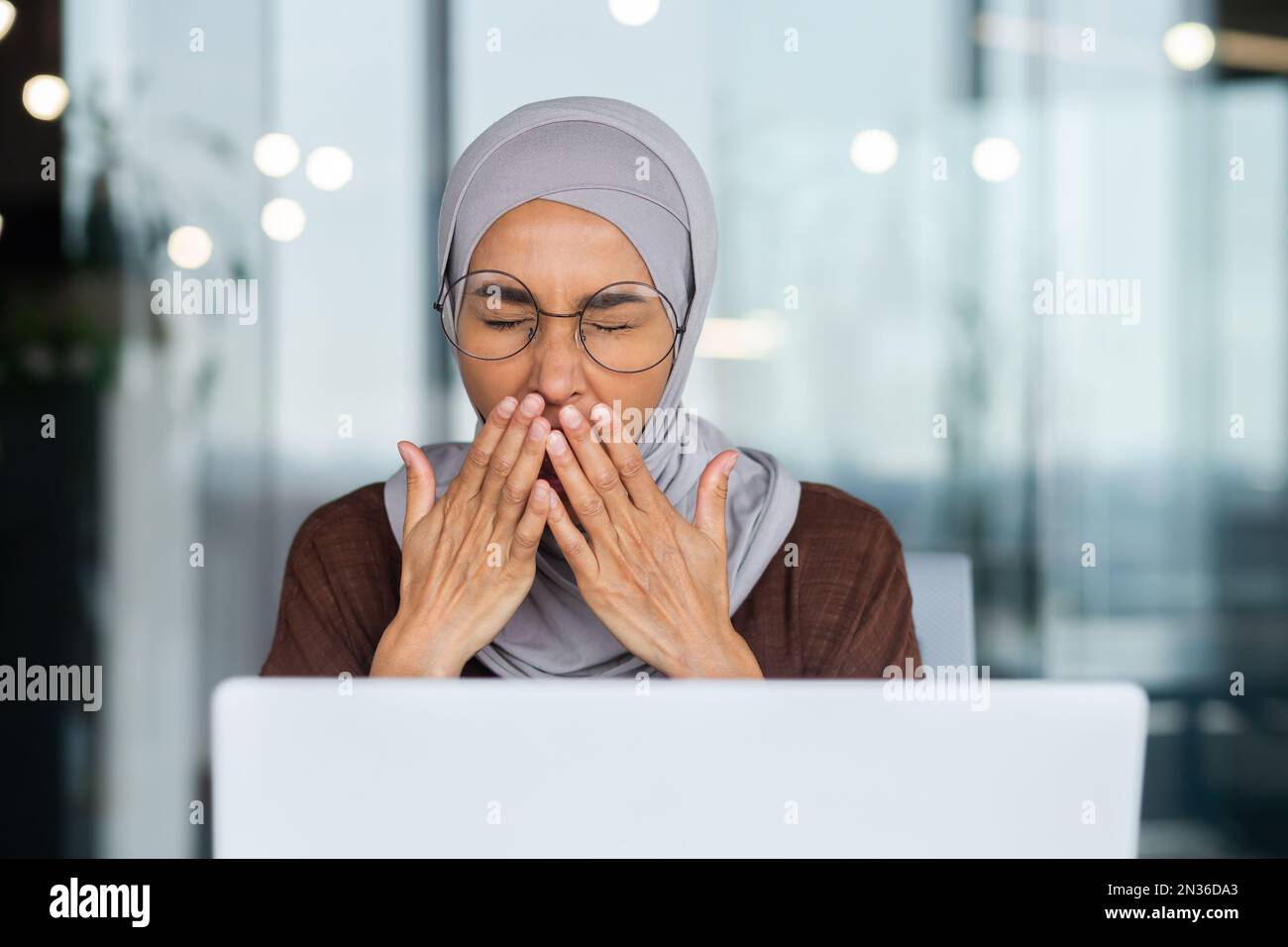 Portrait d'une jeune femme arabe dans le hijab et lunettes assis au bureau  au bureau avec un ordinateur portable. Fatiguée, travail ennuyeux, bâches,  couvre sa bouche avec sa main Photo Stock -