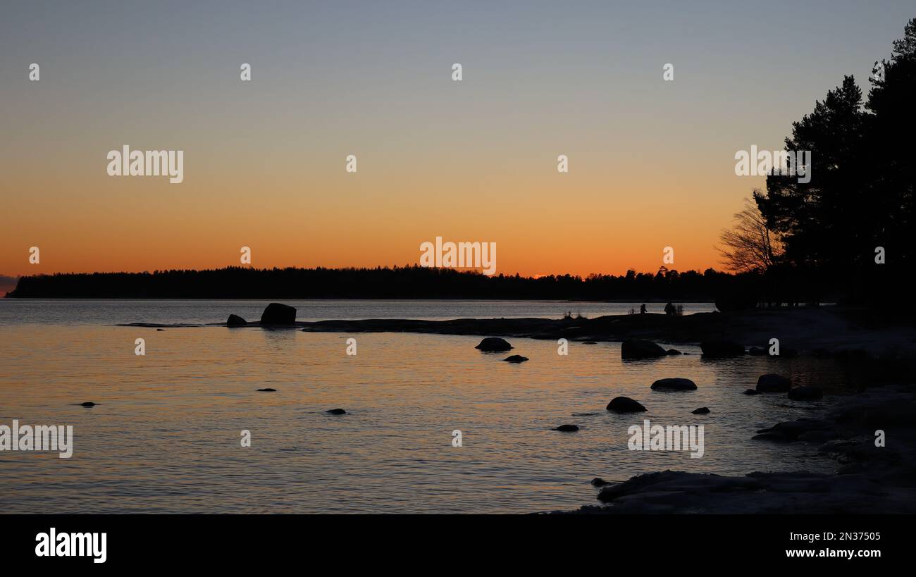 Coucher de soleil rouge au-dessus de la forêt. Mer Baltique avec rochers dans l'ombre. Reflet du coucher de soleil dans la mer. Silhouettes humaines en arrière-plan. Soirée en hiver. Banque D'Images