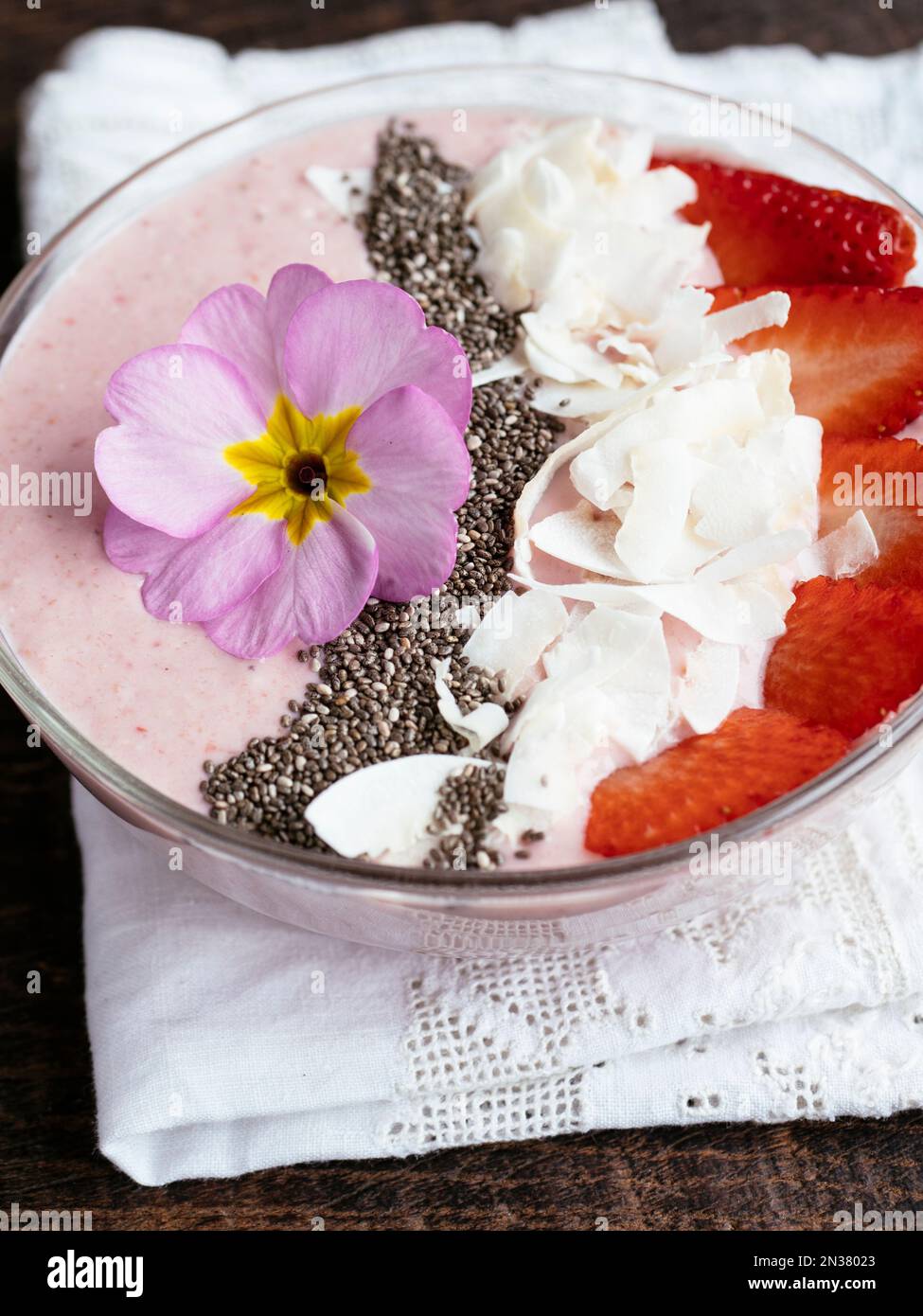 Bol à smoothie au yogourt aux bananes et aux fraises, garni de fraises en tranches, de graines de chia, de flocons de noix de coco et de fleurs comestibles de primula Banque D'Images