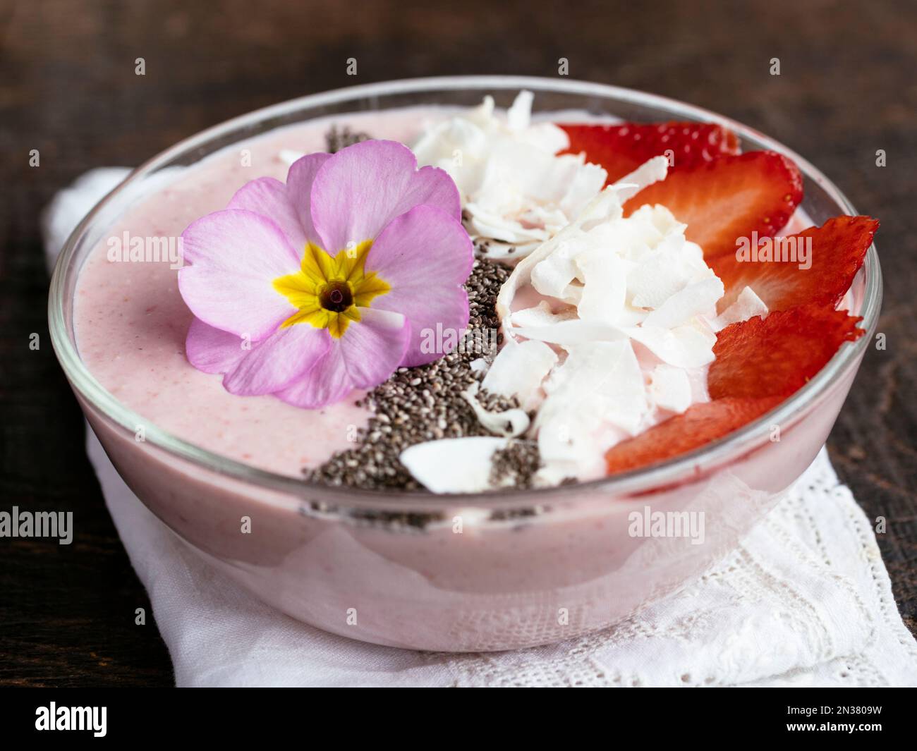 Bol à smoothie au yogourt aux bananes et aux fraises, garni de fraises en tranches, de graines de chia, de flocons de noix de coco et de fleurs comestibles de primula Banque D'Images