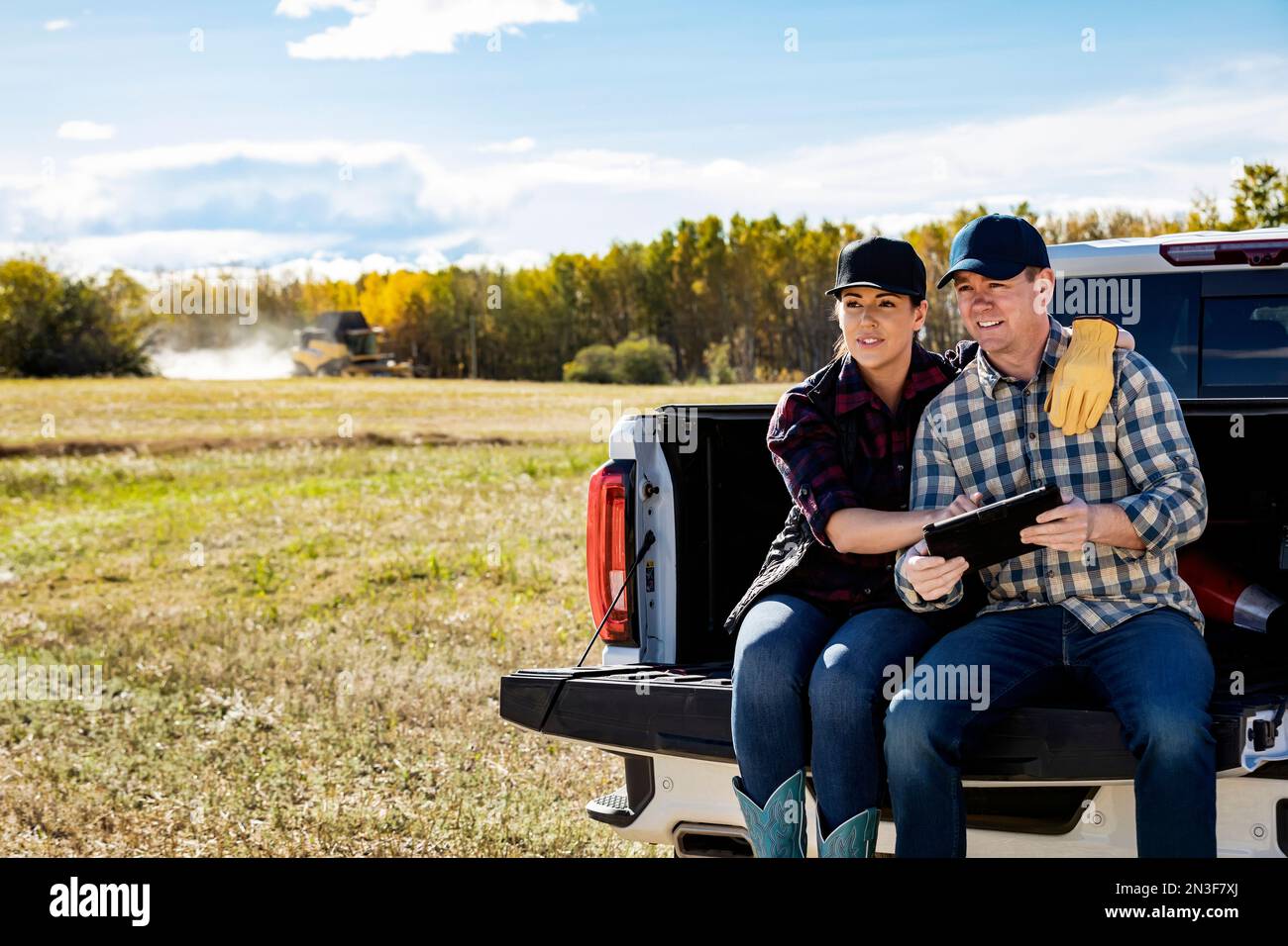 Un mari et sa femme utilisant un appareil portable sans fil pour gérer et surveiller leur récolte de canola assis sur le hayon de leur camion, avec un... Banque D'Images