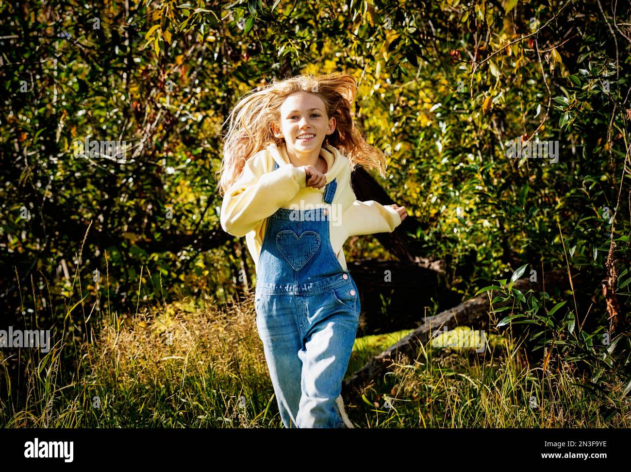 Jeune fille avec de longs cheveux blonds courant hors de la forêt vers la caméra lors d'une chaude journée d'automne dans un parc de la ville ; tenue Albert, Alberta, Canada Banque D'Images