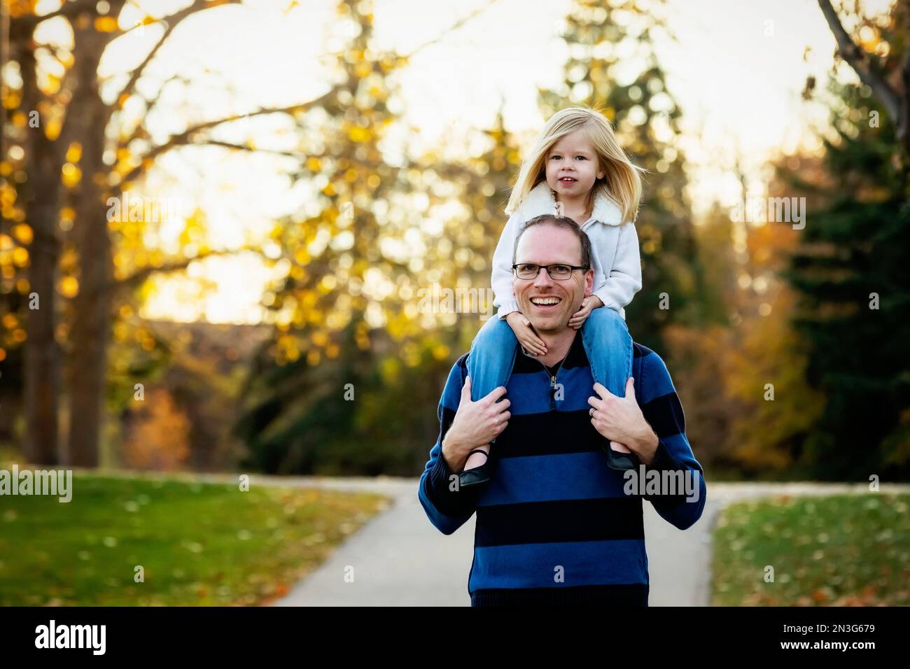 Un père portant sa jeune fille sur ses épaules tout en marchant dans un parc de la ville pendant la saison d'automne; St. Albert, Alberta, Canada Banque D'Images