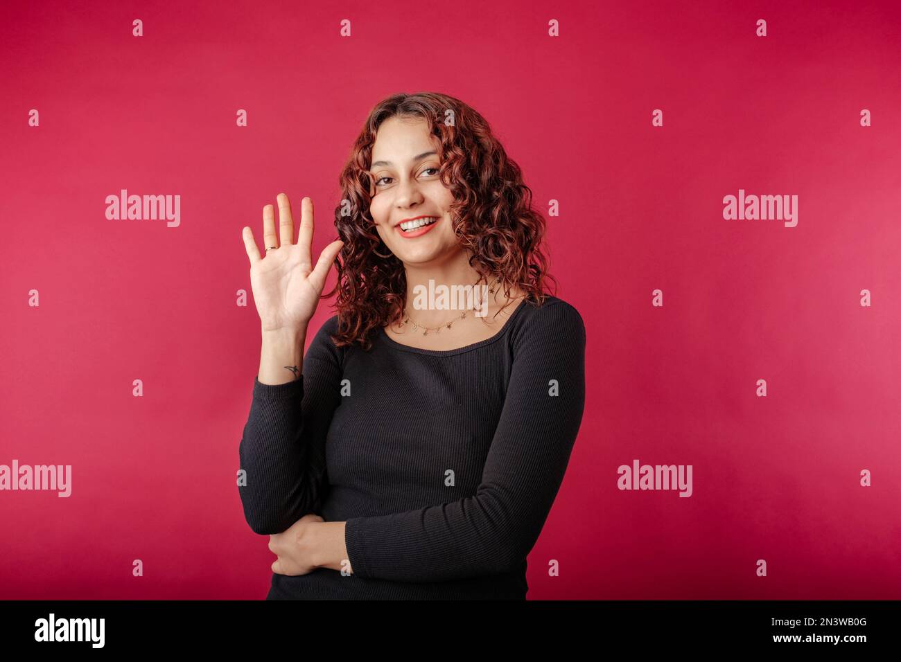 Heureuse femme à tête rouge portant une robe à côtes noires debout isolée sur fond rouge souriant largement tout en agitant la paume relevée, saluant l'ami. Dire Banque D'Images