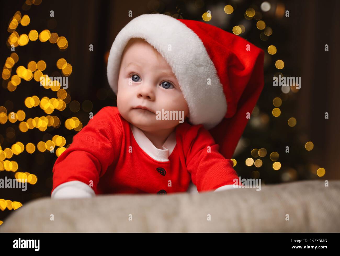 Joli petit bébé en costume de Père Noël contre les lumières de fête floues. Fête de Noël Banque D'Images