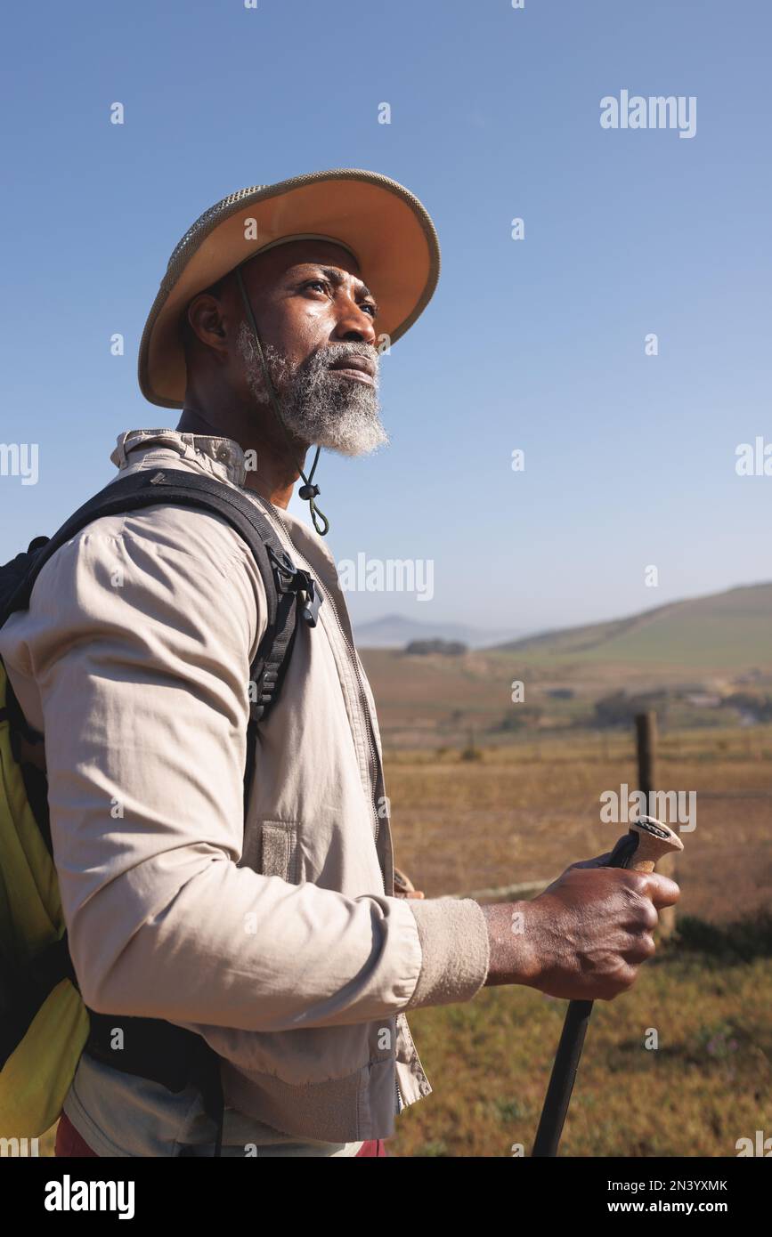 Homme senior afro-américain barbu portant un chapeau avec des bâtons de randonnée debout contre un ciel bleu clair Banque D'Images