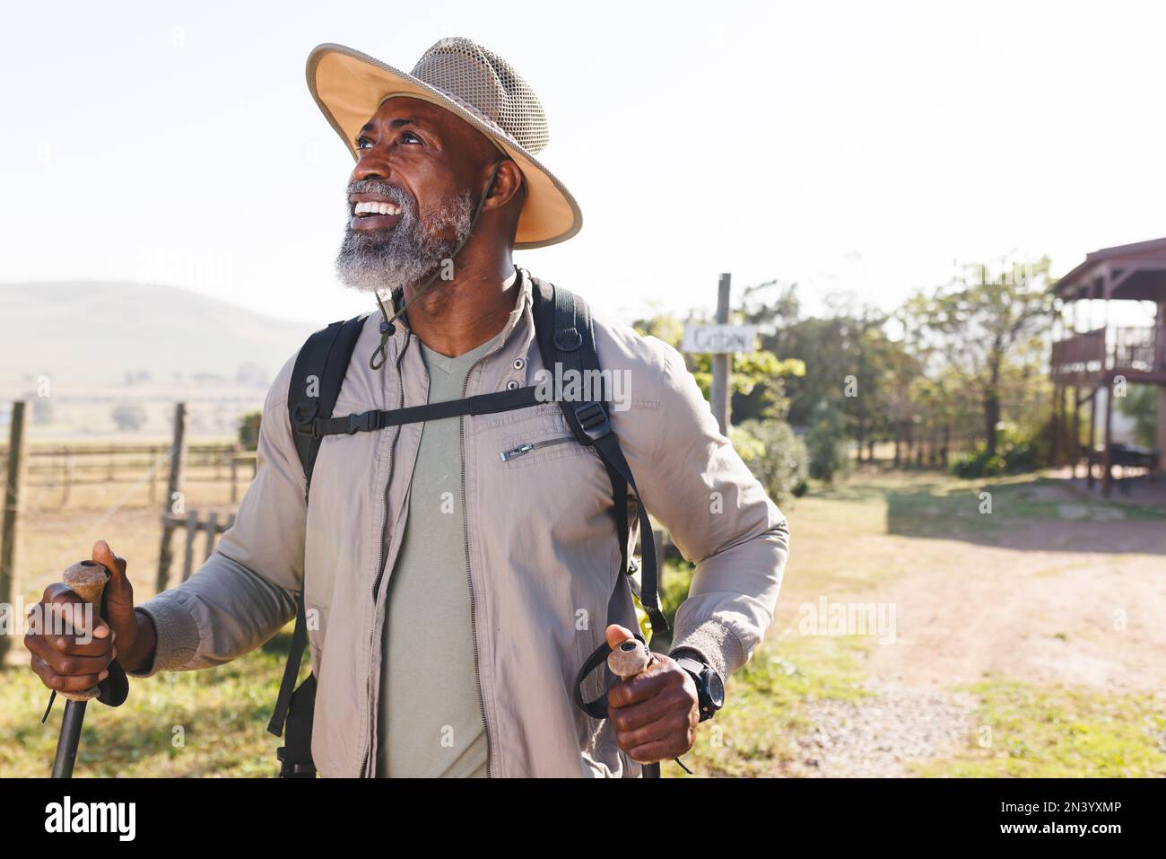 Homme senior afro-américain souriant avec des bâtons de randonnée debout sur terre contre ciel clair Banque D'Images