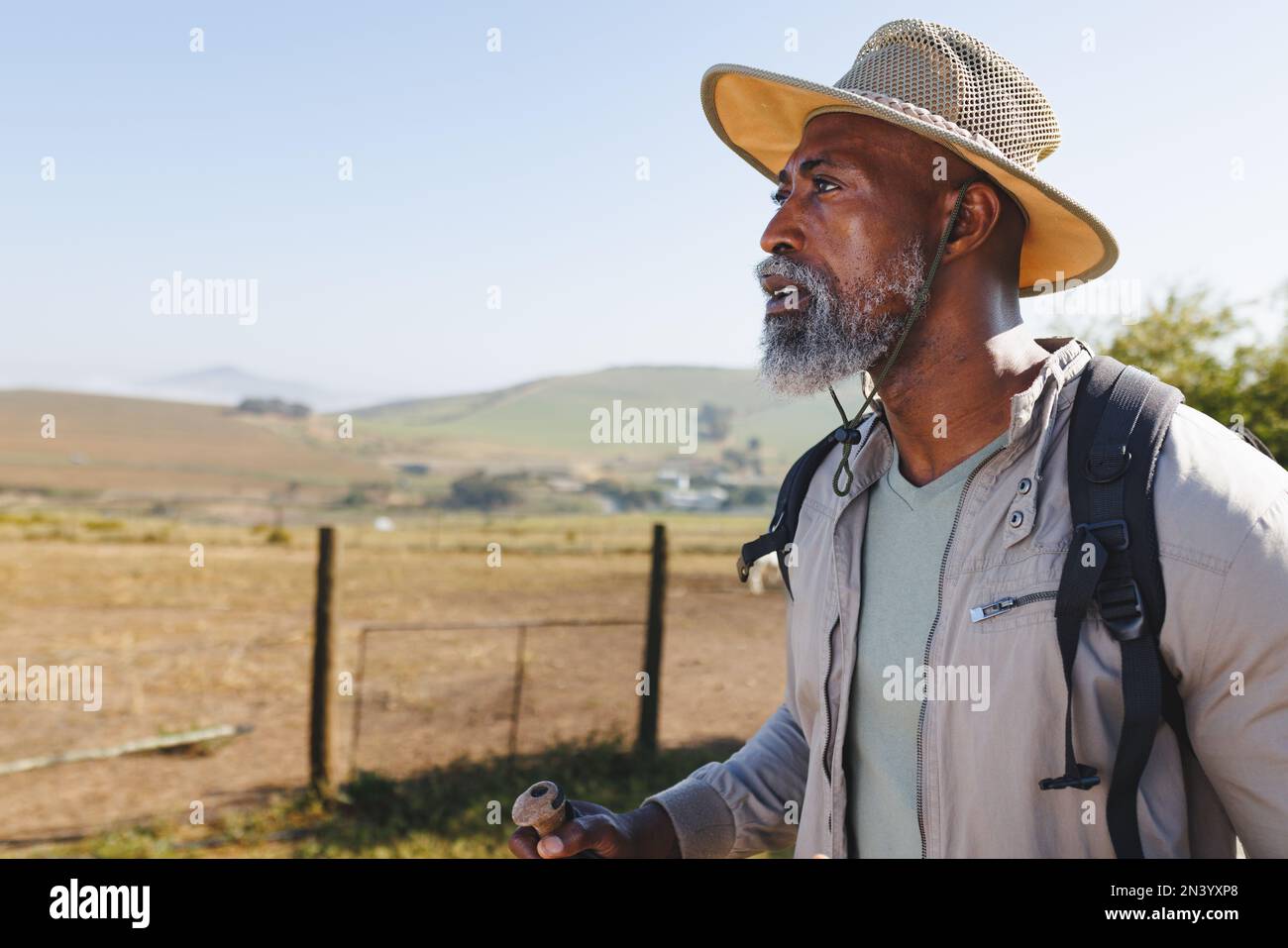 Homme senior afro-américain barbu portant un chapeau et regardant loin tout en randonnée sur terre contre ciel clair Banque D'Images