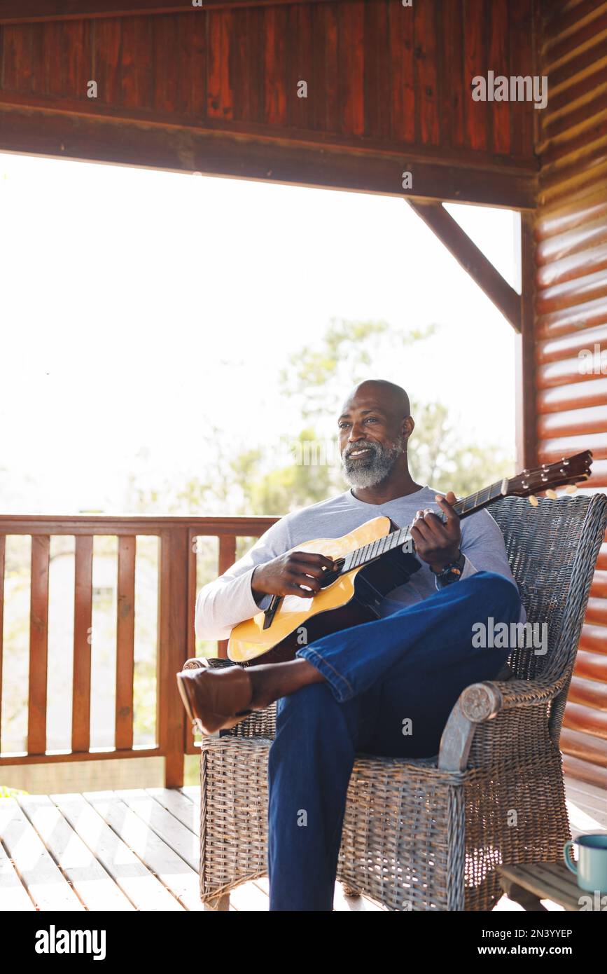 Souriant, homme senior afro-américain, chauve, jouant de la guitare tout en étant assis sur une chaise sur le balcon Banque D'Images