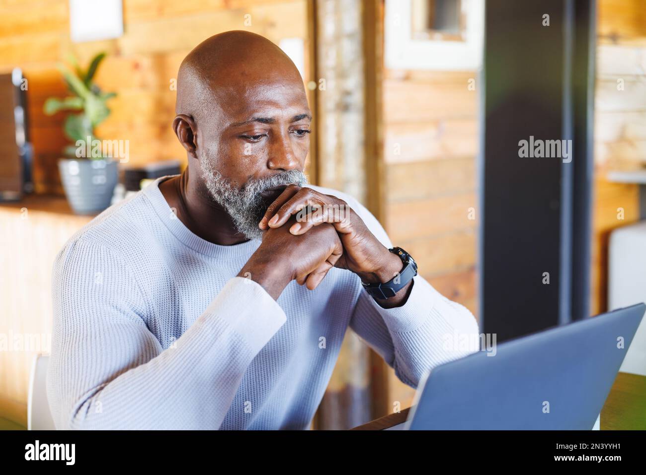 Un homme sérieux et chauve, un homme âgé d'afrique et d'amérique, avec ses mains jointes, regarde un ordinateur portable dans une cabine en rondins Banque D'Images