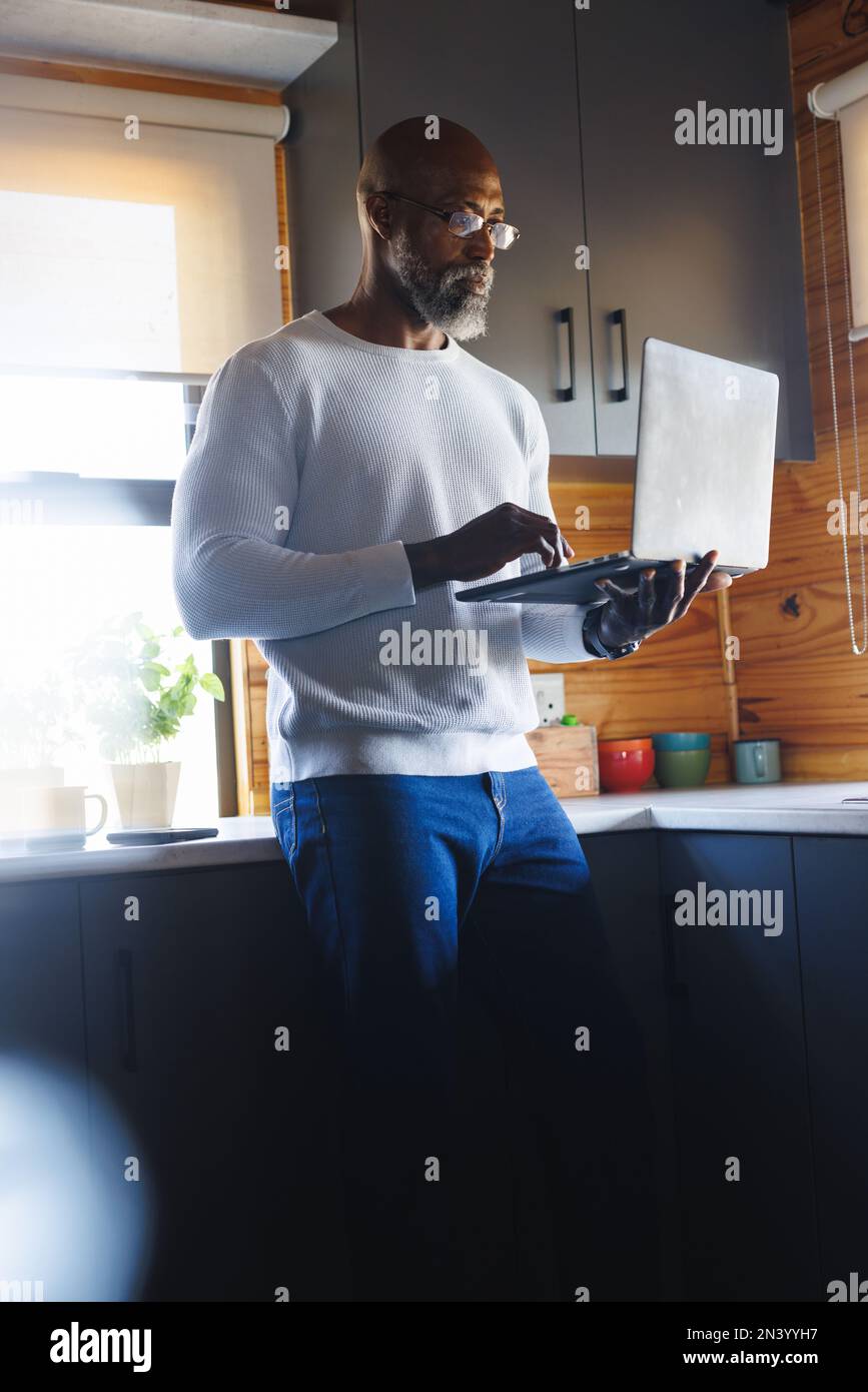 Grave homme d'amérique chauve senior portant des lunettes en utilisant un ordinateur portable en se tenant dans la cuisine Banque D'Images