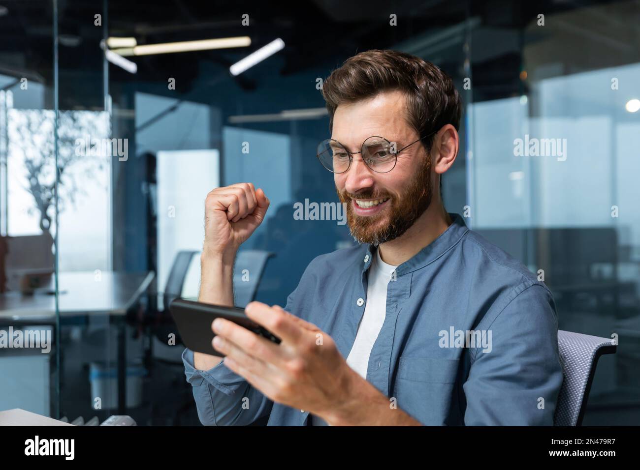 Homme au bureau sur le lieu de travail jouant à des jeux vidéo sur smartphone, homme d'affaires heureux de gagner se réjouit et tient la main dans le geste de triomphe, patron dans le maillot heureux de la réussite et du résultat. Banque D'Images