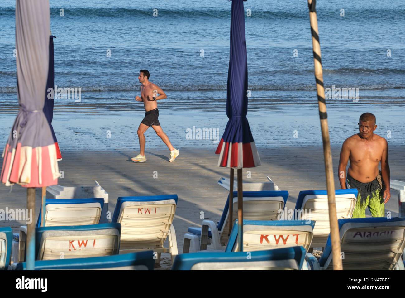 En début de matinée, un touriste occidental se lance sur la plage de Kamala à Phuket, en Thaïlande, tandis qu'un homme local organise des chaises longues et des parasols de plage Banque D'Images