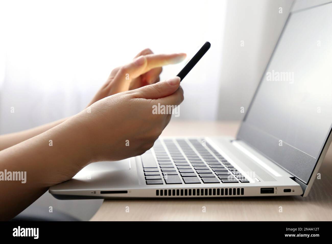 Mains féminines avec smartphone sur l'arrière-plan du clavier de l'ordinateur portable. Femme avec téléphone mobile assis près de la fenêtre, concept de communication en ligne Banque D'Images