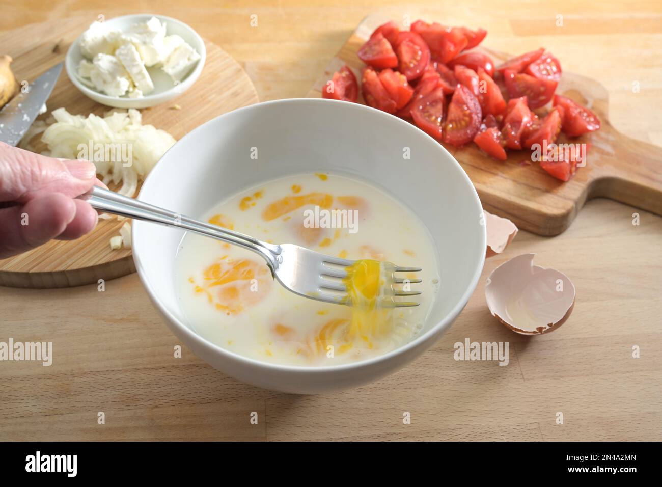 Les oeufs et le lait sont mélangés avec une fourchette dans un bol blanc sur un plan de cuisine en bois, préparation de la cuisson pour une omelette ou frittata avec des tomates et des oignons, Banque D'Images