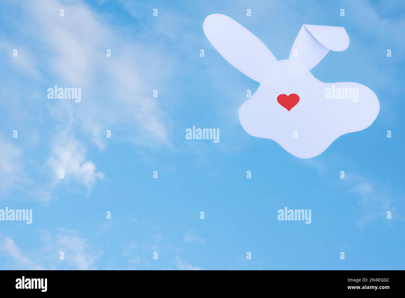 Carte créative composée d'une silhouette de lapin en papier blanc et d'un cœur rouge au lieu d'un nez sur fond ciel nuageux. Concept de vacances de printemps minimales Banque D'Images