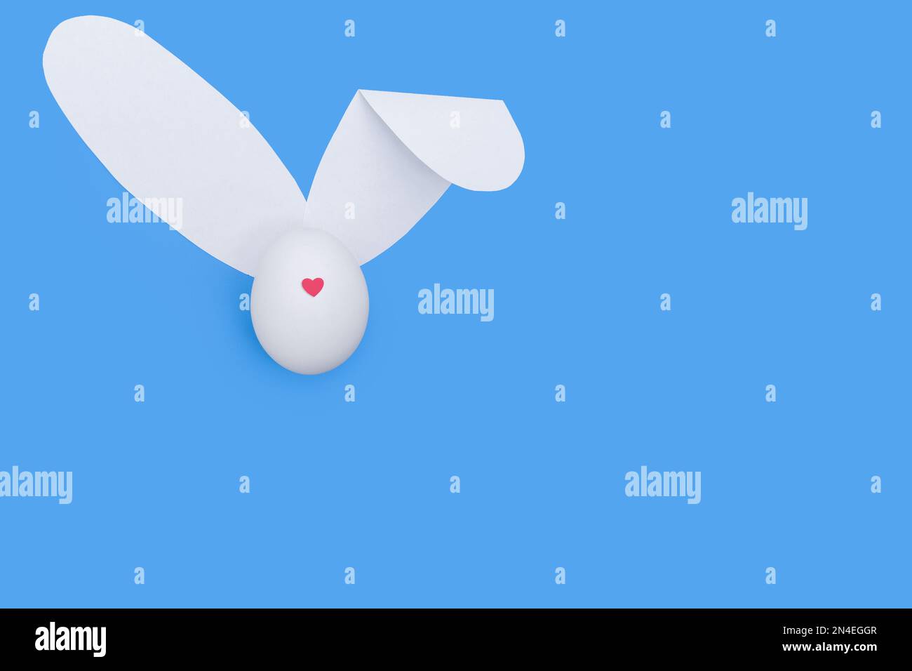 Une carte de vœux créative faite d'un œuf blanc avec un cœur rouge au lieu d'un nez et d'oreilles de papier blanc comme un lapin sur fond bleu ciel. Le minimum Banque D'Images
