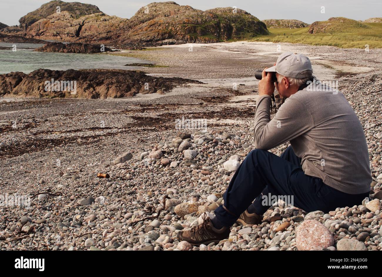 Man 60+ assis sur une plage de pierres Camas Cuil et t-Saimh, sur le côté ouest d'Iona, en Écosse, en regardant la mer à travers des jumelles Banque D'Images