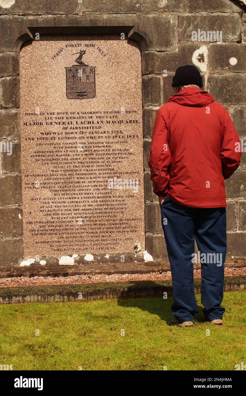 Un homme 60+ debout, un manteau rouge et un chapeau noir, lisant une plaque de pierre à l'extrémité du mausolée de Macquarie sur le domaine de Gruline, Mull, Écosse Banque D'Images