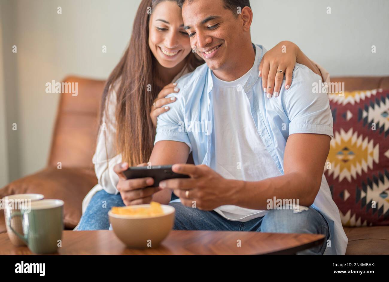 Un jeune couple souriant s'embrasse tout en regardant un smartphone. Couple multiethnique partageant les réseaux sociaux sur smartphone. Fille africaine souriante embrassant fro Banque D'Images