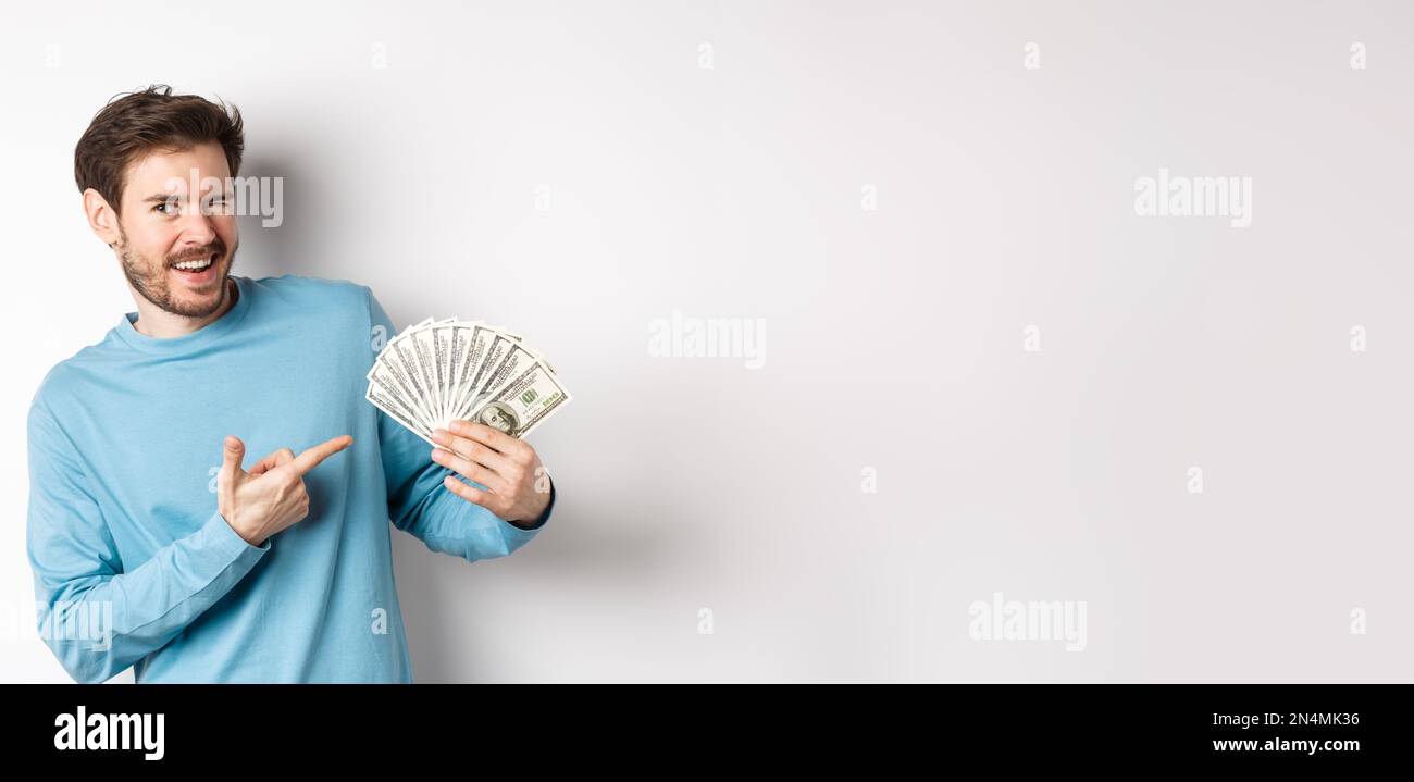 Beau gars riche qui se vante à la caméra, montrant des revenus, pointant le doigt sur l'argent gagné et souriant, debout sur fond blanc Banque D'Images