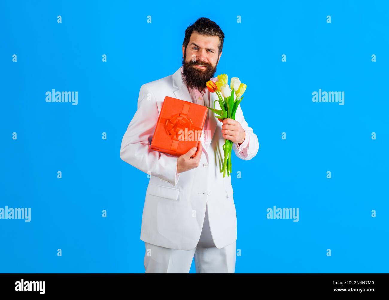 Homme barbu souriant avec boîte et bouquet de fleurs. Homme d'affaires en costume avec cadeau et bouquet de tulipes. Boîte de présentation. Fleur de printemps. Cadeau pour Banque D'Images