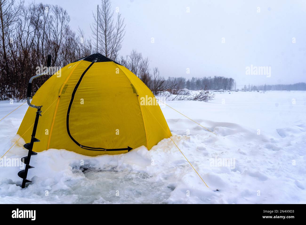 Une tente de pêche jaune d'hiver se trouve à côté d'un bloc de glace sur la neige et la glace dans un Blizzard sur le lac près des arbres. Banque D'Images