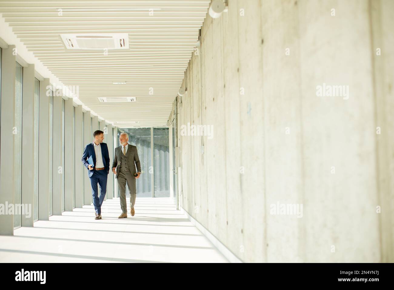 Un jeune homme d'affaires et un homme d'affaires senior descendent dans le couloir d'un bureau, profondément dans la conversation. Ils sont tous deux habillés professionnellement, reflétant leur acum d'affaires Banque D'Images