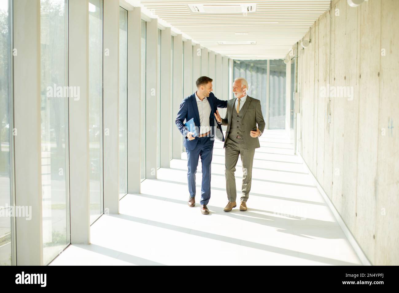 Un jeune homme d'affaires et un homme d'affaires senior descendent dans le couloir d'un bureau, profondément dans la conversation. Ils sont tous deux habillés professionnellement, reflétant leur acum d'affaires Banque D'Images