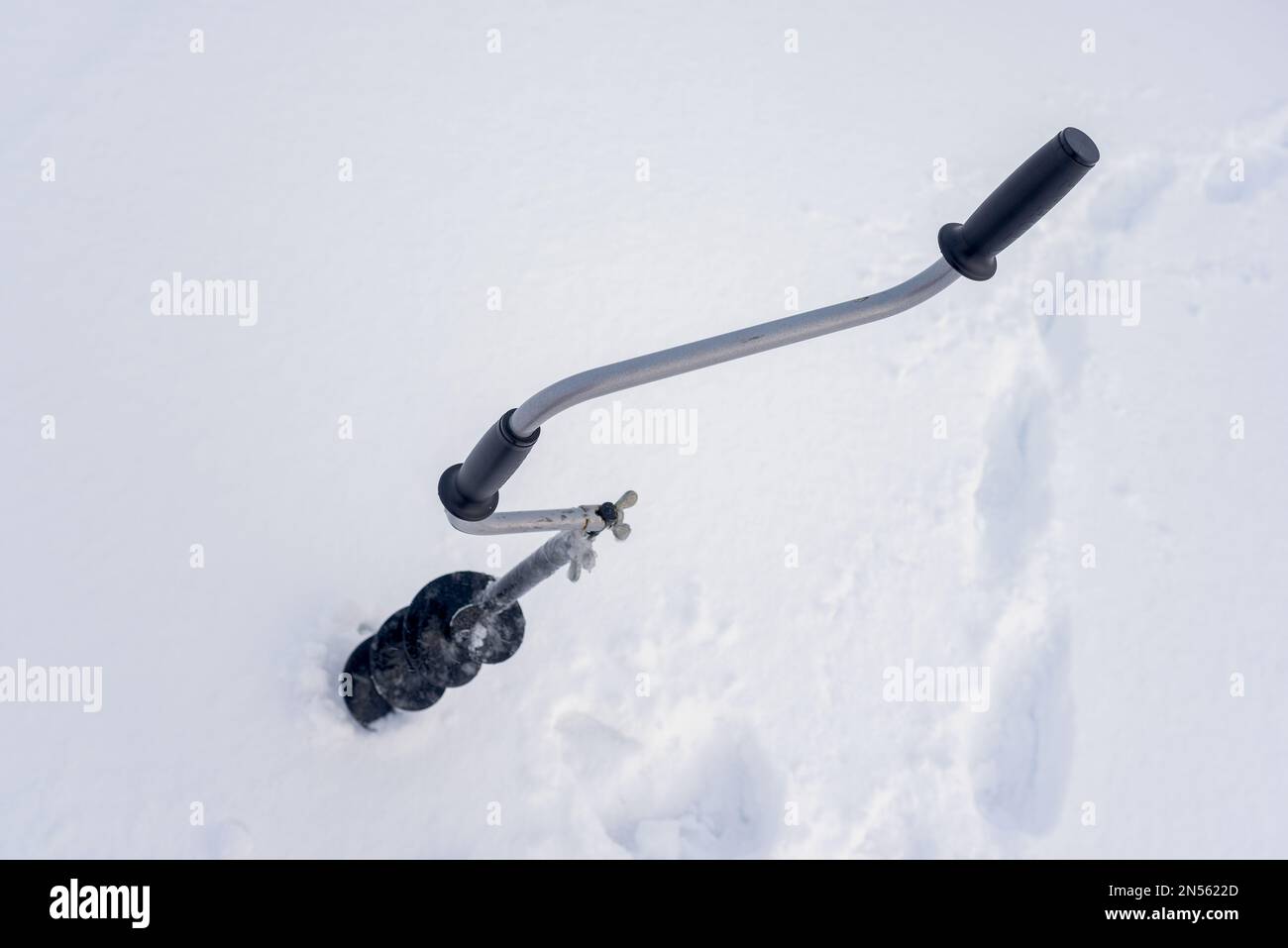 Une perceuse à glace à main noire en fer avec poignée incurvée se visse dans la glace en hiver sur fond de neige blanche. Banque D'Images