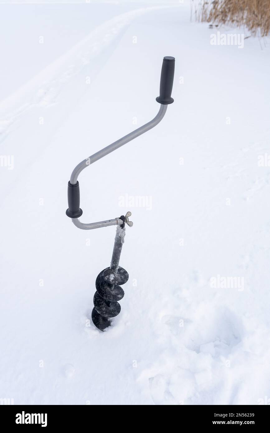 Une perceuse à glace à main noire en fer avec une poignée incurvée et des vis congelées dans de la glace est vissée dans la glace en hiver sur fond blanc Banque D'Images