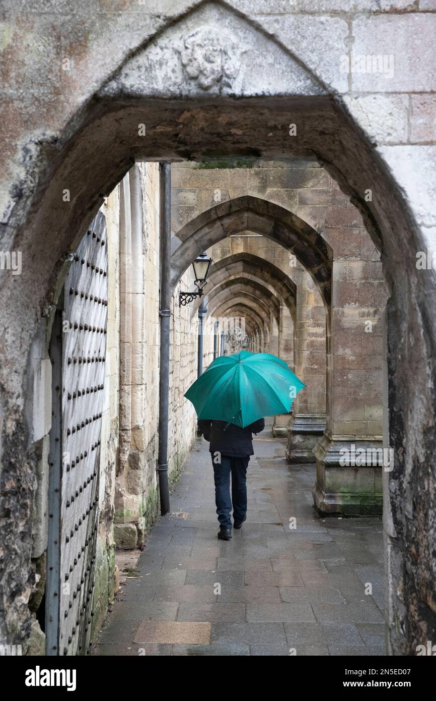 Homme marchant sous la pluie avec un parapluie sous les arches des contreforts volants sur le côté de la cathédrale de Winchester, Winchester, Hampshire, Angleterre, Royaume-Uni Banque D'Images