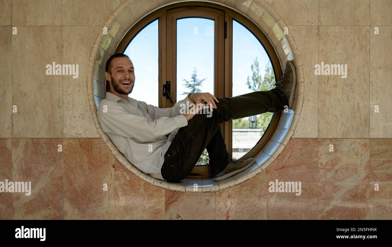 Attrayant jeune homme blanc pose pour la caméra dans une fenêtre circulaire Banque D'Images