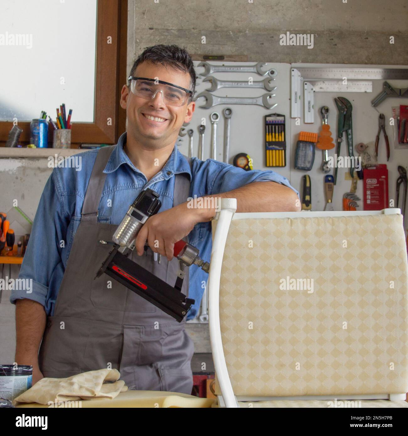 Un tapisseur souriant dans son atelier avec divers outils et un pistolet à ongles après avoir restauré une vieille chaise. Banque D'Images