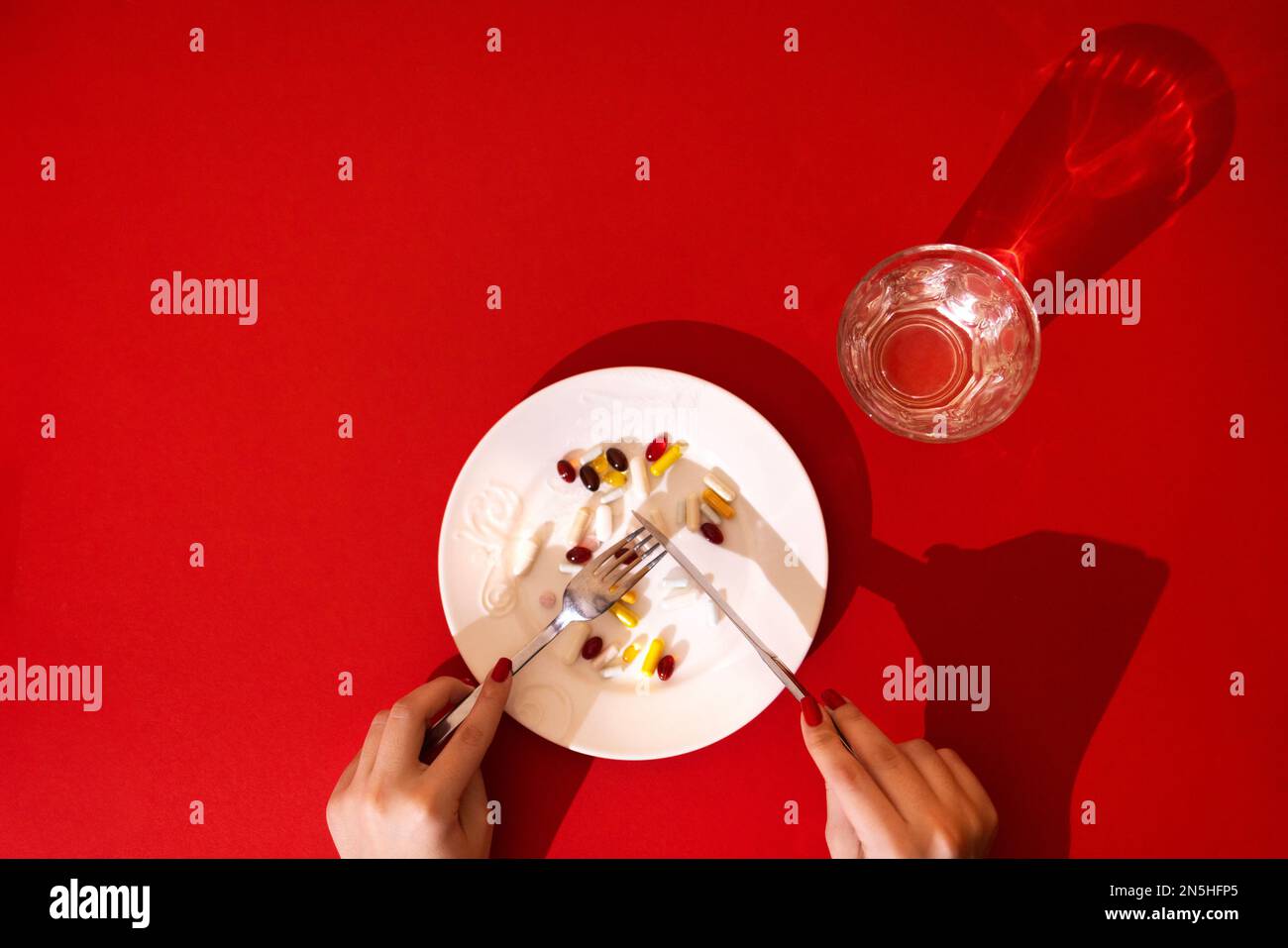 Comprimés, pilules et capsules sur une plaque en porcelaine blanche. La table est équipée d'une plaque, d'une fourchette, d'un couteau et d'un verre à eau. Mains de la femme sur rouge vif b Banque D'Images