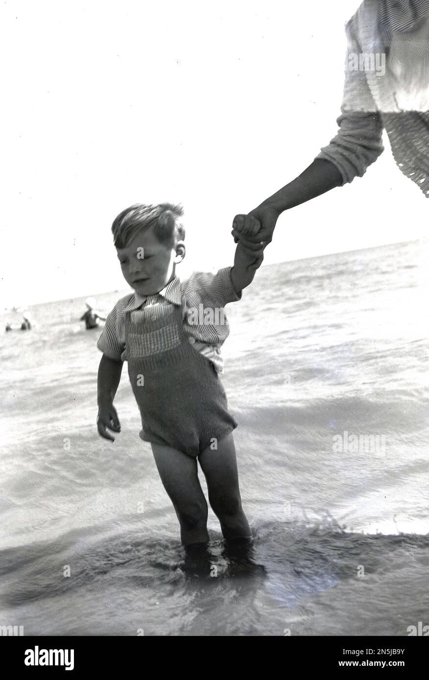 Années 1950, historique, main aidante.... une mère tenant la main d'un petit garçon portant un maillot de bain en laine, car il a un padde dans la mer, Angleterre, Royaume-Uni. Banque D'Images