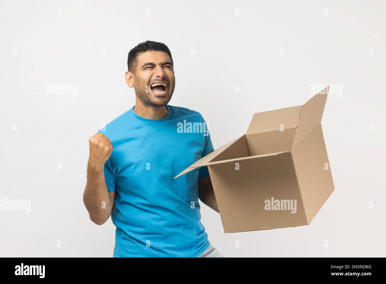 Portrait d'un homme extrêmement heureux non rasé portant un T-shirt bleu debout avec une boîte en carton dans la main, clenches poing, célébrant le succès. Prise de vue en studio isolée sur fond gris. Banque D'Images