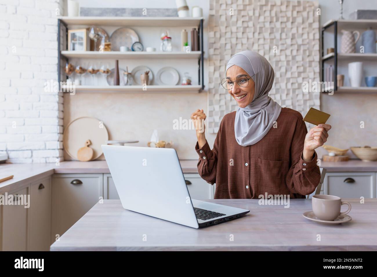 Bonne femme shopper dans la boutique en ligne célébrant la victoire et les réductions d'achat réussies, les ventes, la femme musulmane dans hijab shopping en ligne en utilisant l'ordinateur portable et la carte de crédit bancaire à l'intérieur à la maison dans la cuisine. Banque D'Images