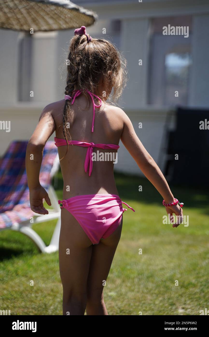 Une fille en maillot de bain rose sur l'herbe près des chaises longues  regarde quelque chose de la vue arrière. L'enfant s'amuse avec les vacances  d'été. Vacances en famille par Photo Stock -