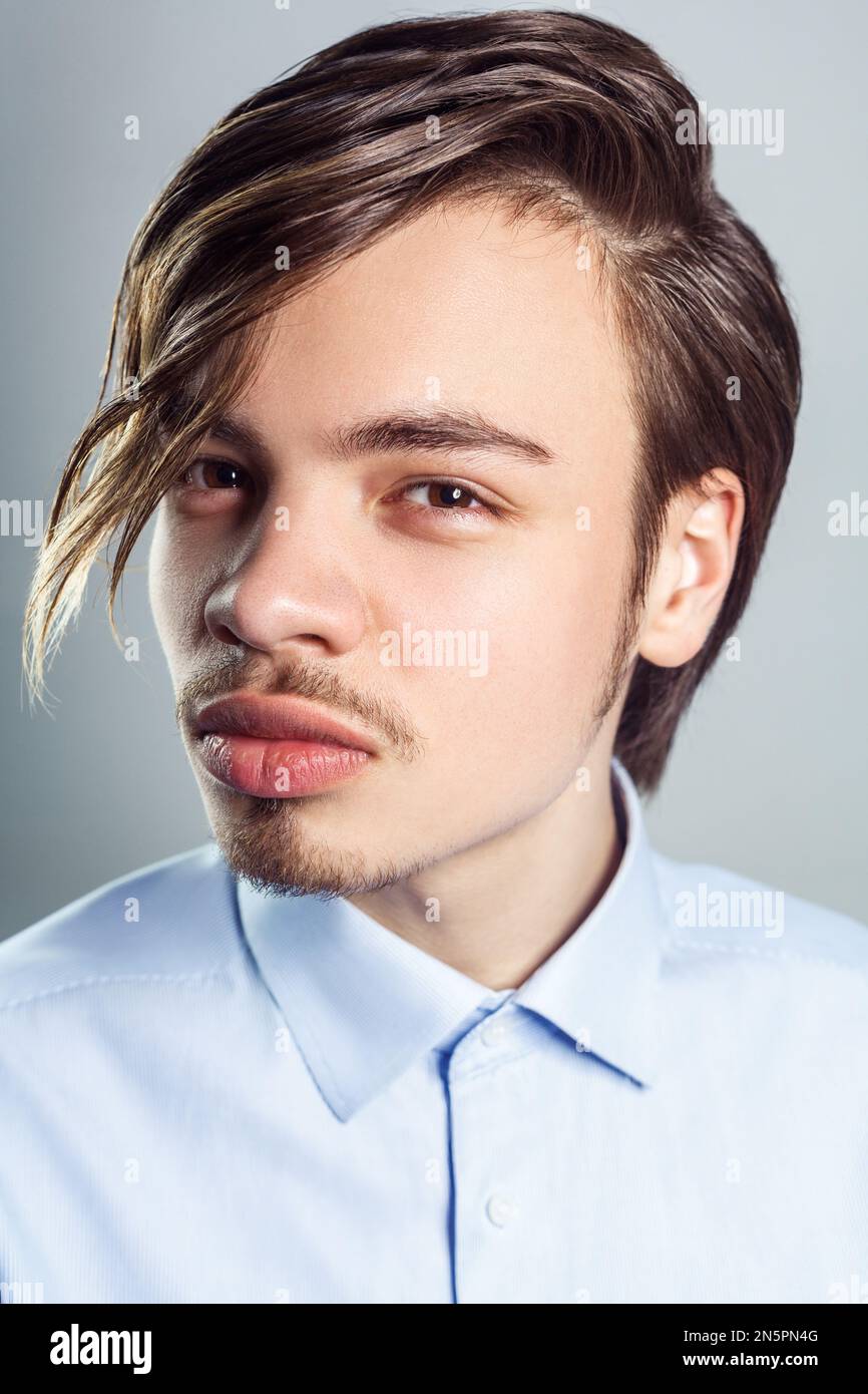 Portrait d'un jeune homme beau avec une longue coiffure à franges sur les yeux, regardant l'appareil photo avec un look mystérieux, portant une chemise bleue. Prise de vue en studio isolée sur fond gris. Banque D'Images
