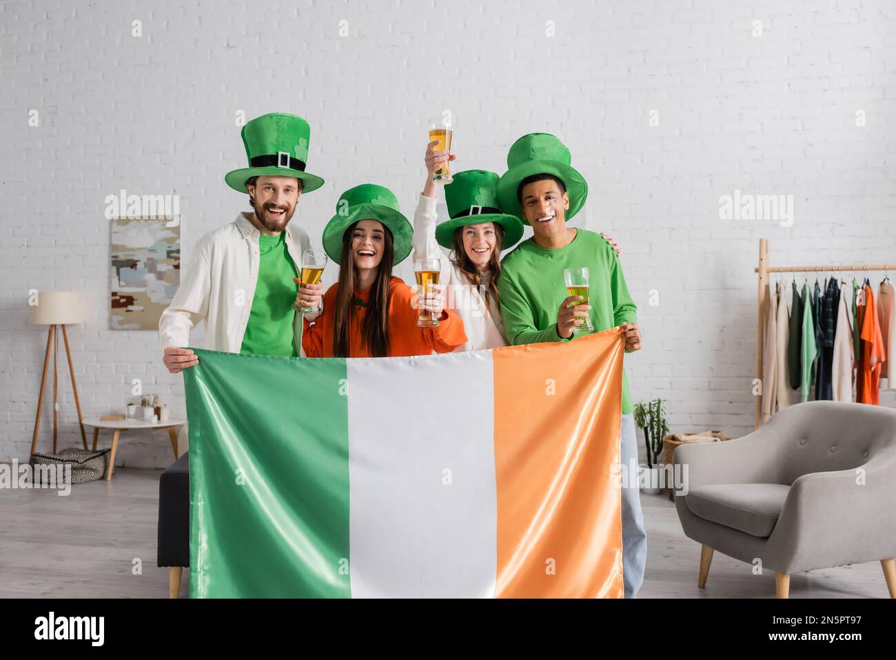 Des amis heureux et multiculturels en chapeaux verts portant des verres de bière et un drapeau irlandais tout en célébrant le jour de Saint Patrick Banque D'Images