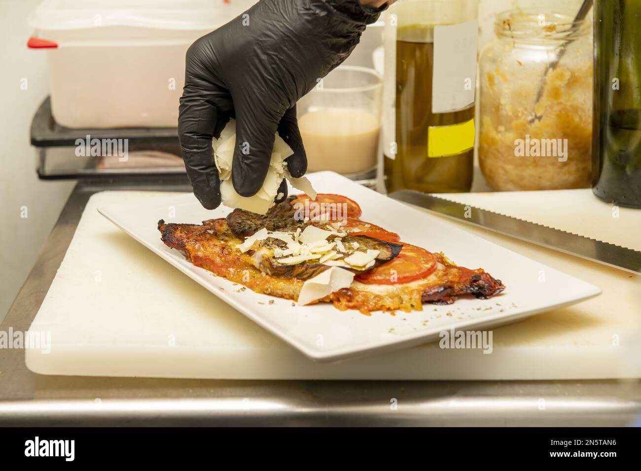 Un chef aux mains gantées de plastique noir mettant les dernières touches de copeaux de parmesan sur un milanais Banque D'Images