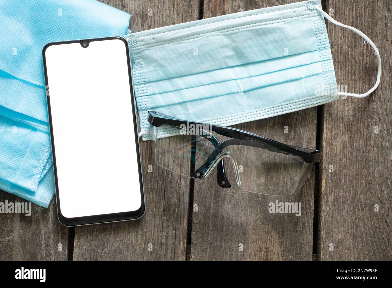 téléphone avec écran blanc vierge à côté des lunettes et un masque médical sur une table en bois Banque D'Images