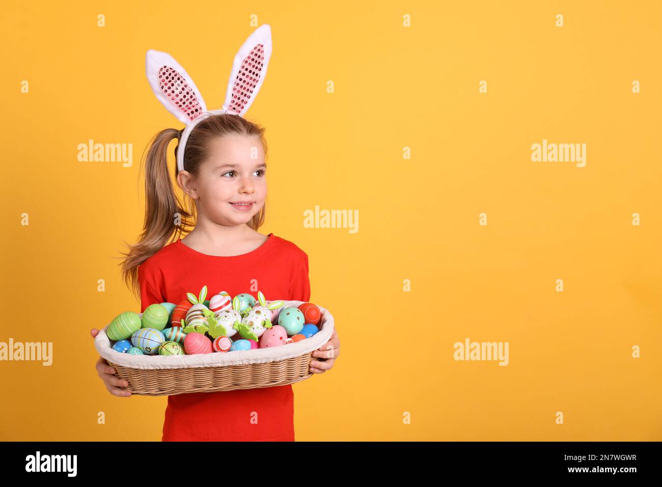 Adorable petite fille aux oreilles de lapin tenant un panier en osier rempli d'œufs de Pâques sur fond orange. Espace pour le texte Banque D'Images