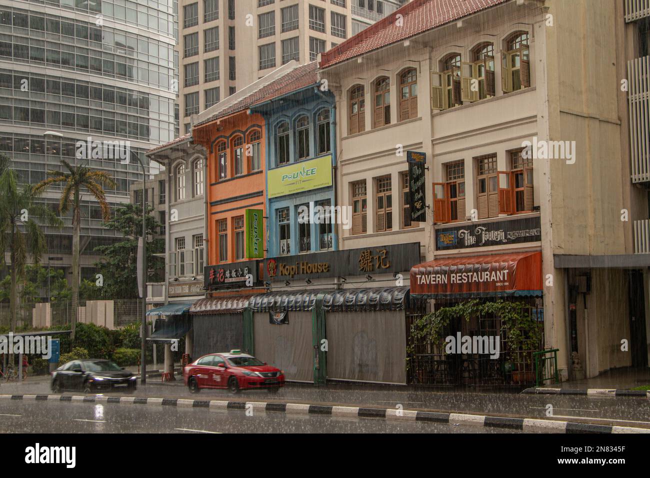 Une rangée de demeures coloniales historiques colorées avec des magasins, des enseignes de restaurants et des voitures qui descendent dans la rue le long d'eux lors d'une journée de pluie à Singapour. Banque D'Images